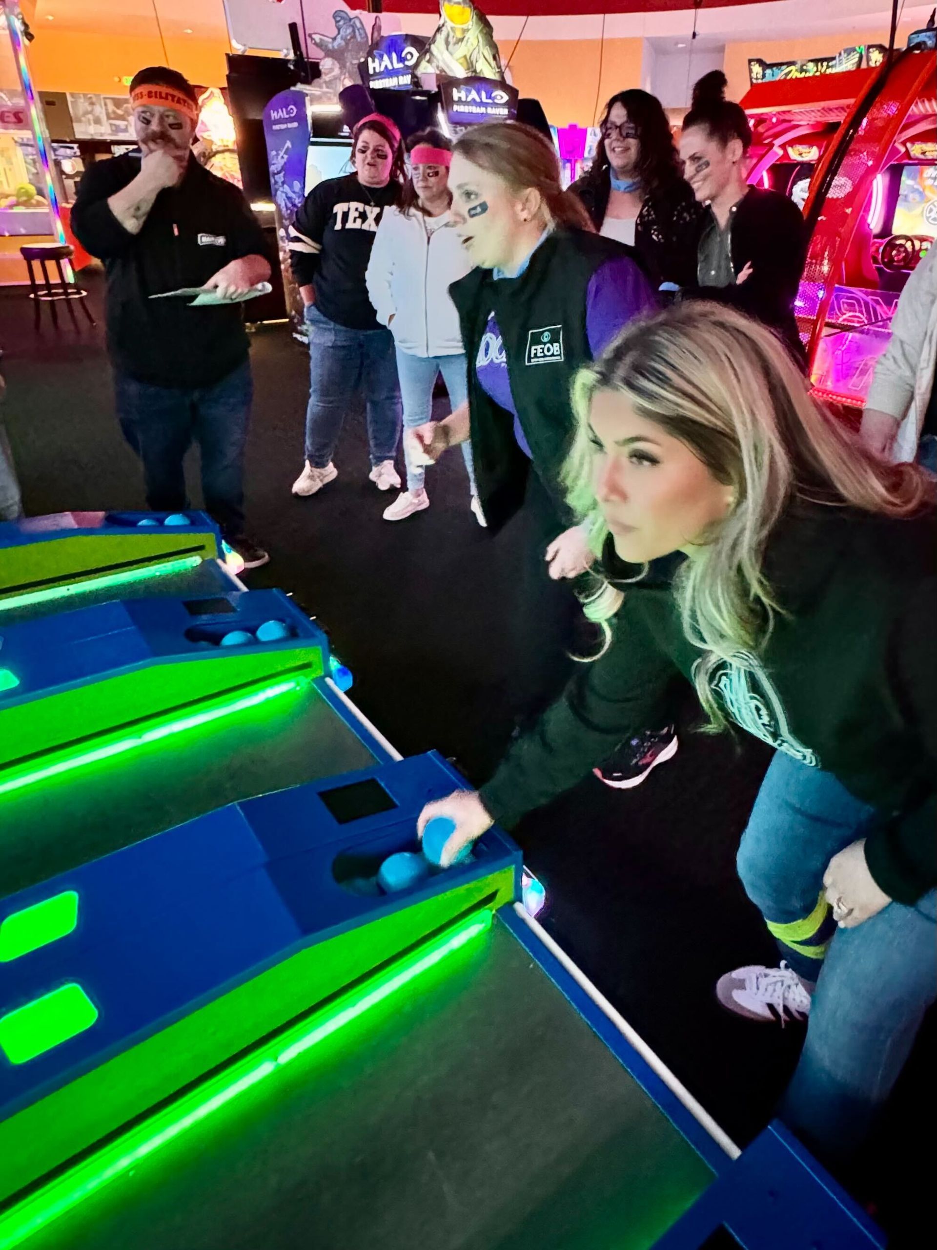 People playing an arcade game. Woman focuses, rolling a ball, others watch. Bright lights, indoors.