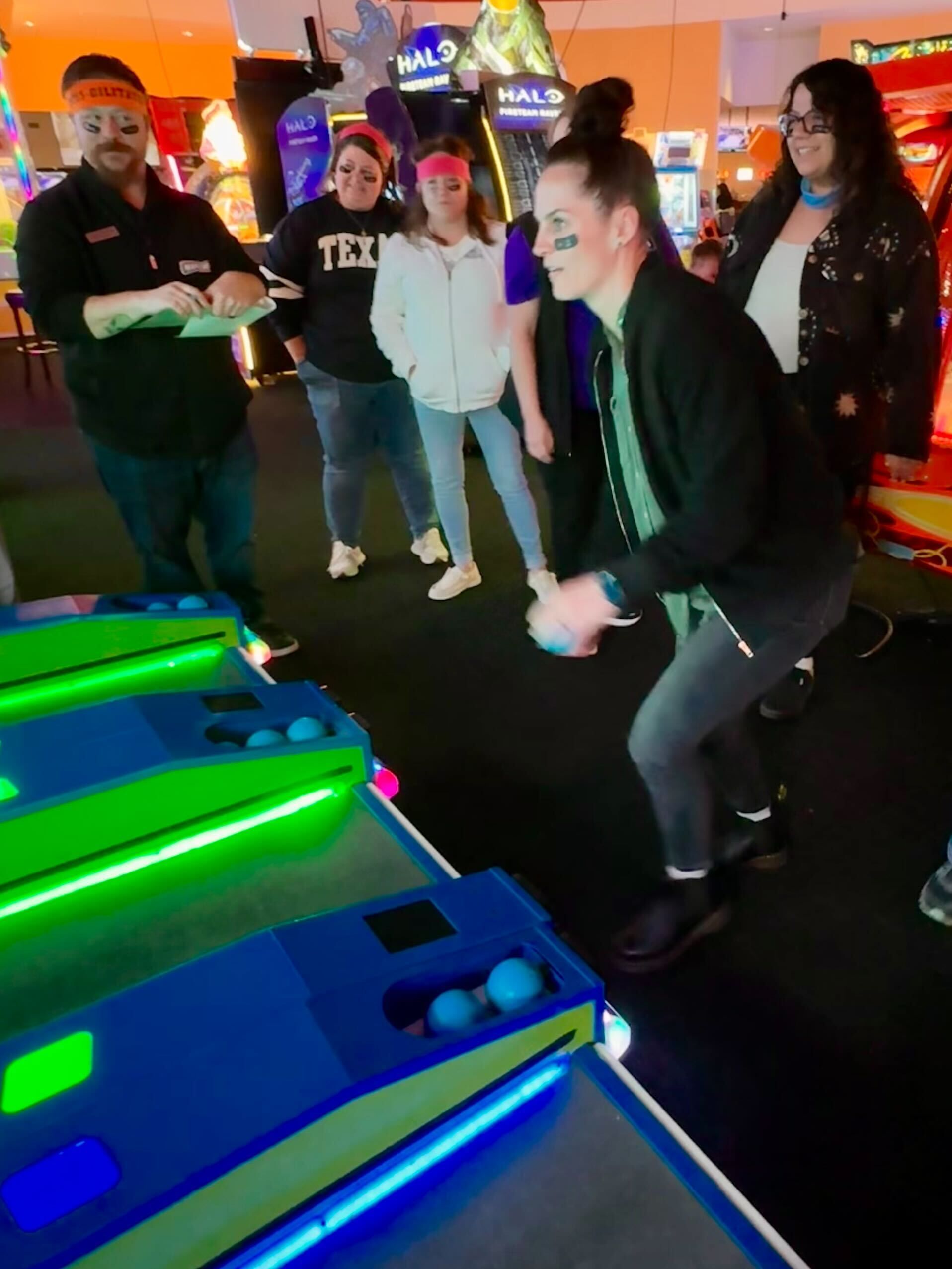 People playing an arcade game; woman throws a ball at the target. Others watch, some with headbands, in an arcade.