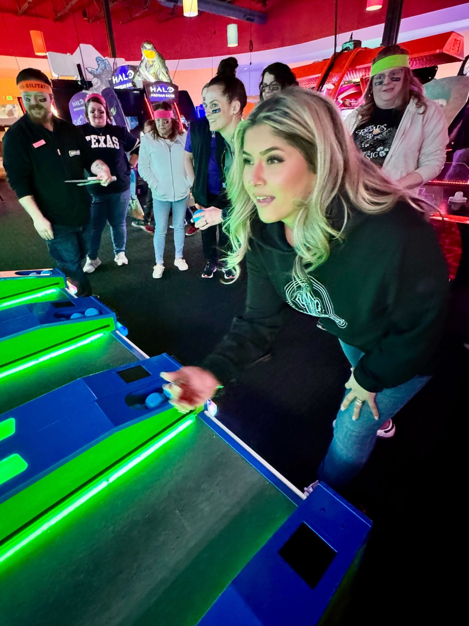 Woman playing arcade game, others watch. Bright neon lights, woman with focused expression.