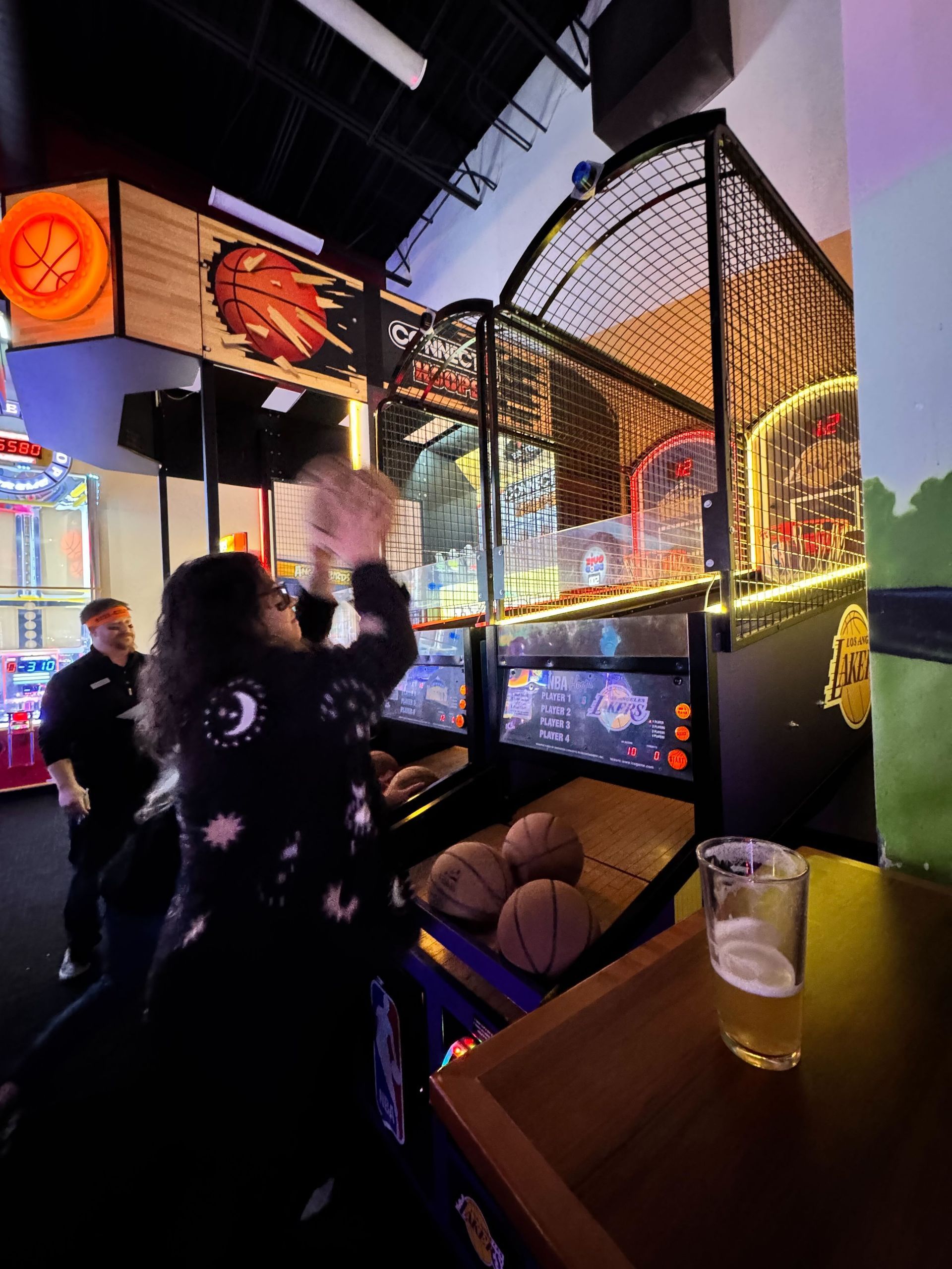 Person playing a basketball arcade game, holding a beer. Indoor arcade setting.