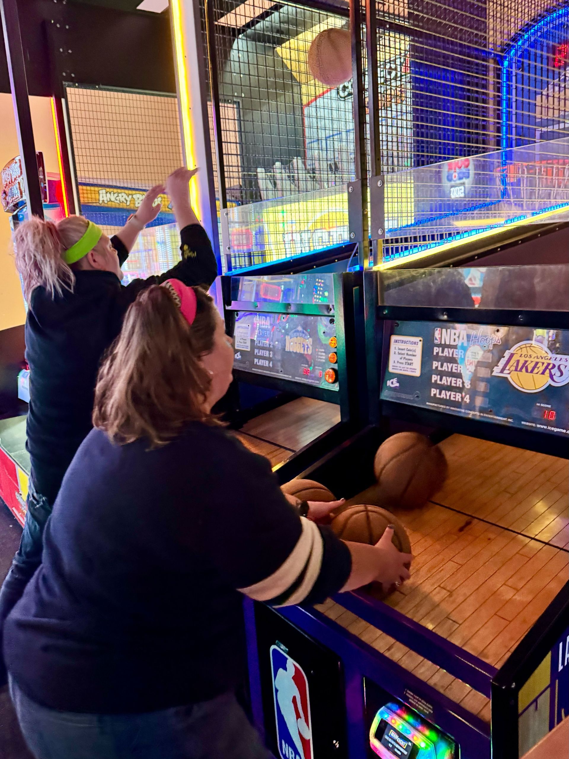 Two women playing a basketball arcade game. One shoots a ball; the other holds more. Inside an arcade.