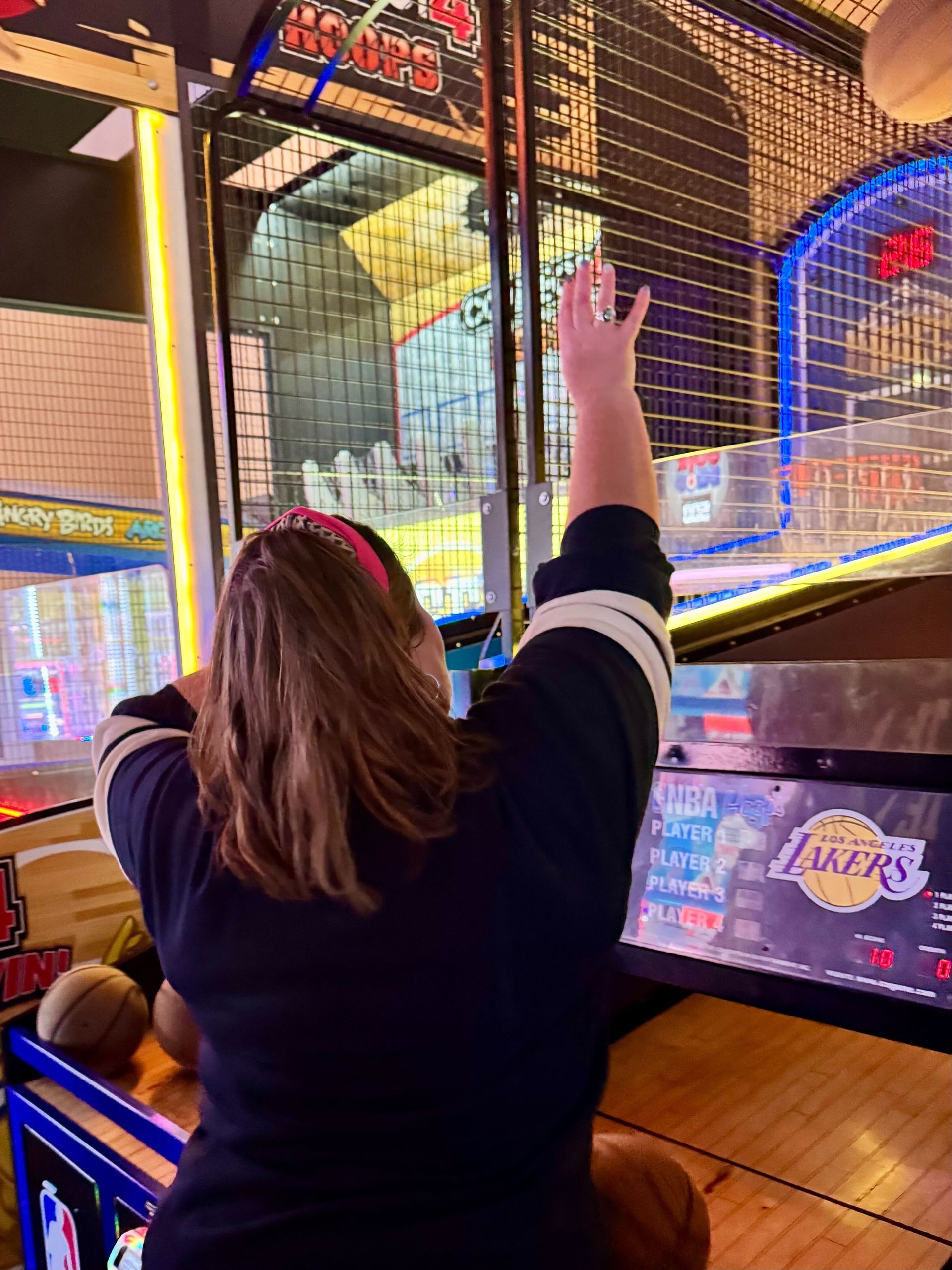 Woman playing basketball arcade game, reaching arm up to shoot. Lakers logo visible.