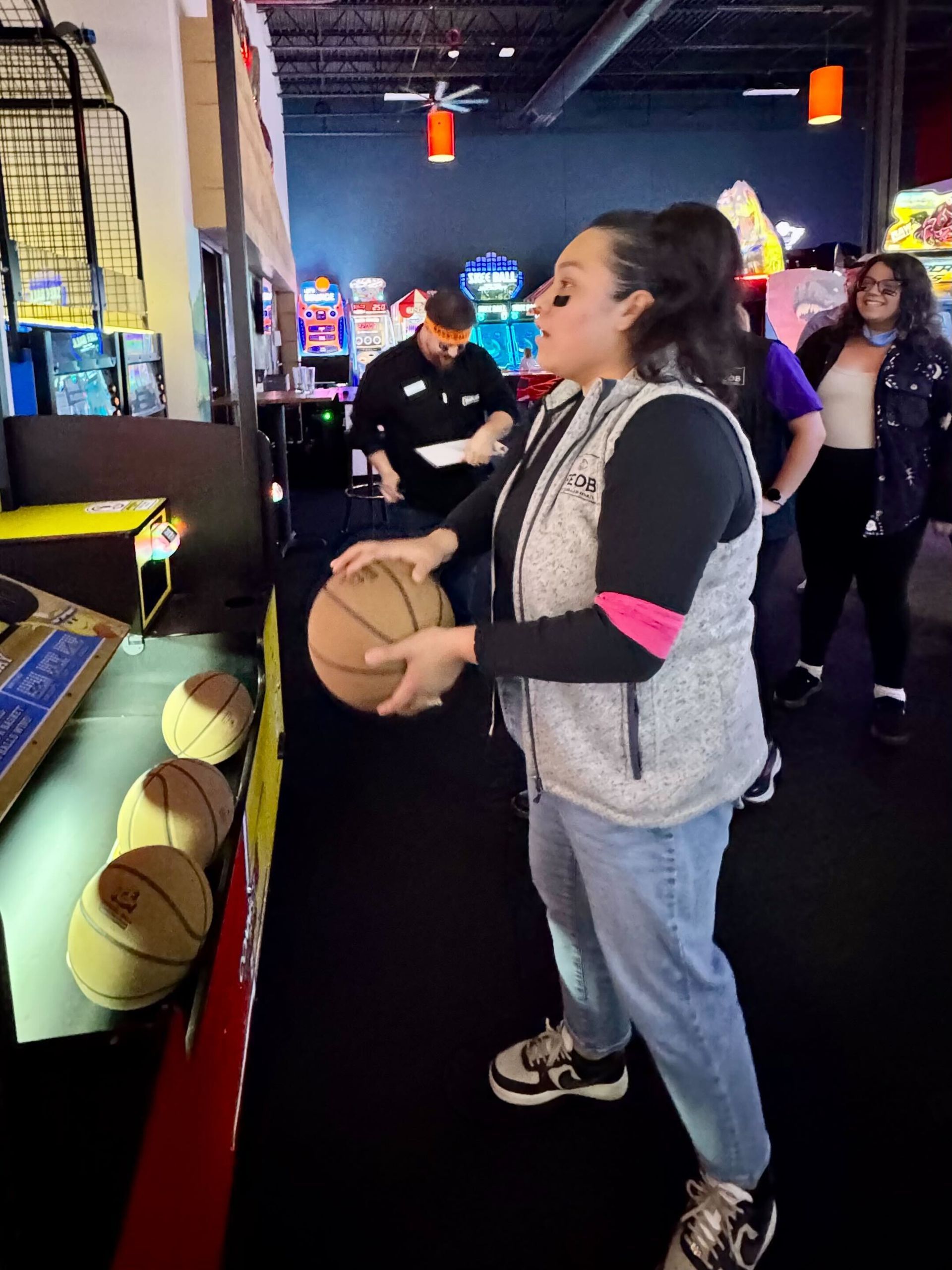 Woman shooting a basketball at an arcade game, holding the ball, wearing a vest and jeans. Others watch.