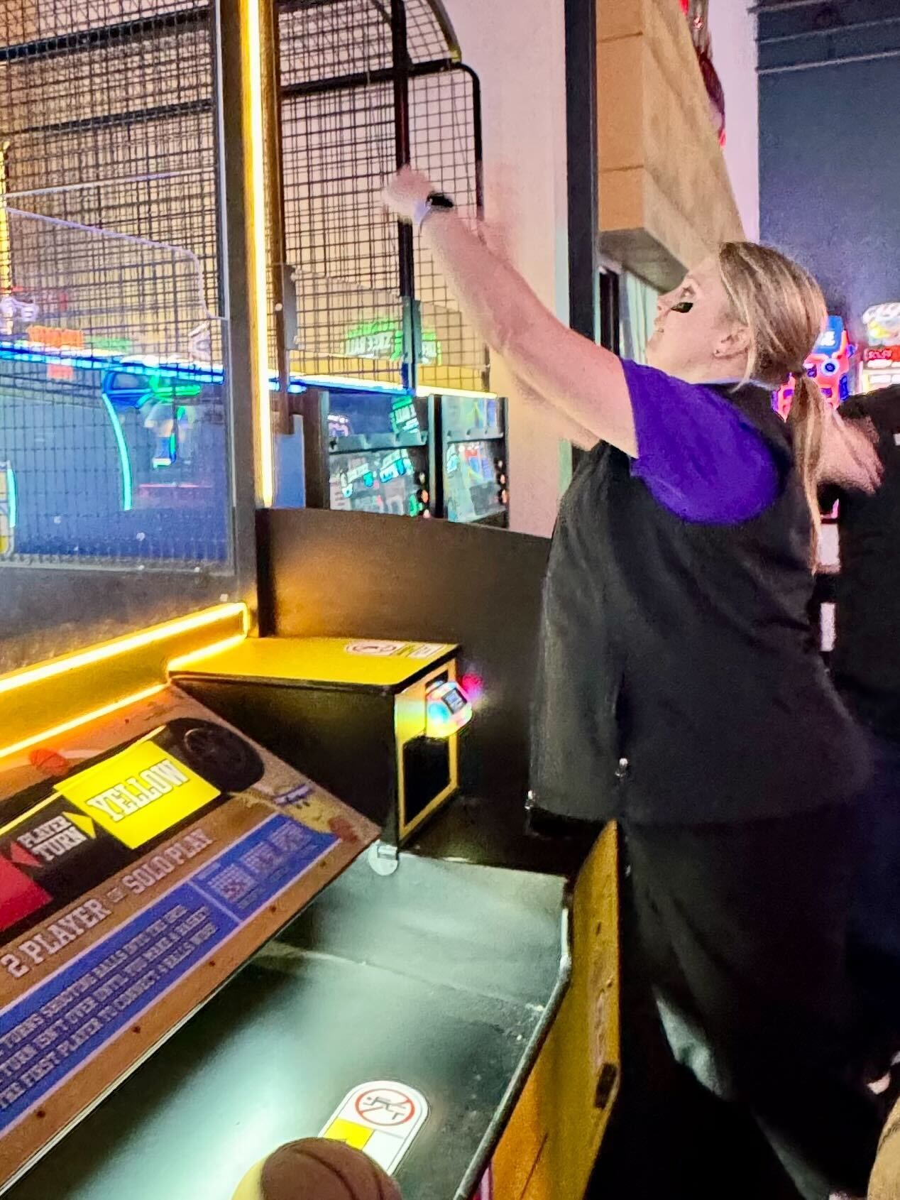 Woman playing basketball arcade game, reaching up to shoot. Purple shirt, black vest, indoor setting.