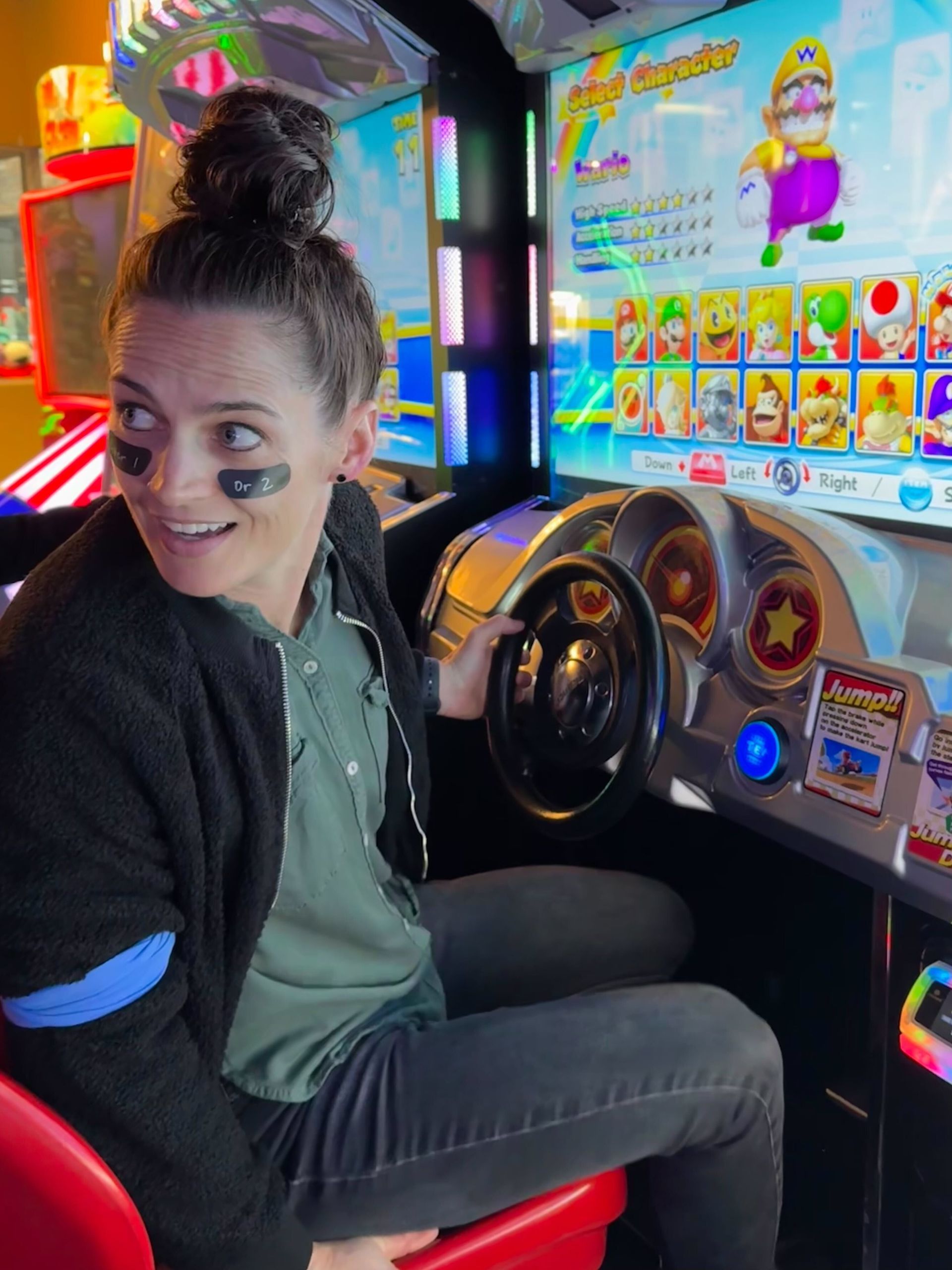 Woman with a bun playing Mario Kart arcade game, looking to the left with eye black on.