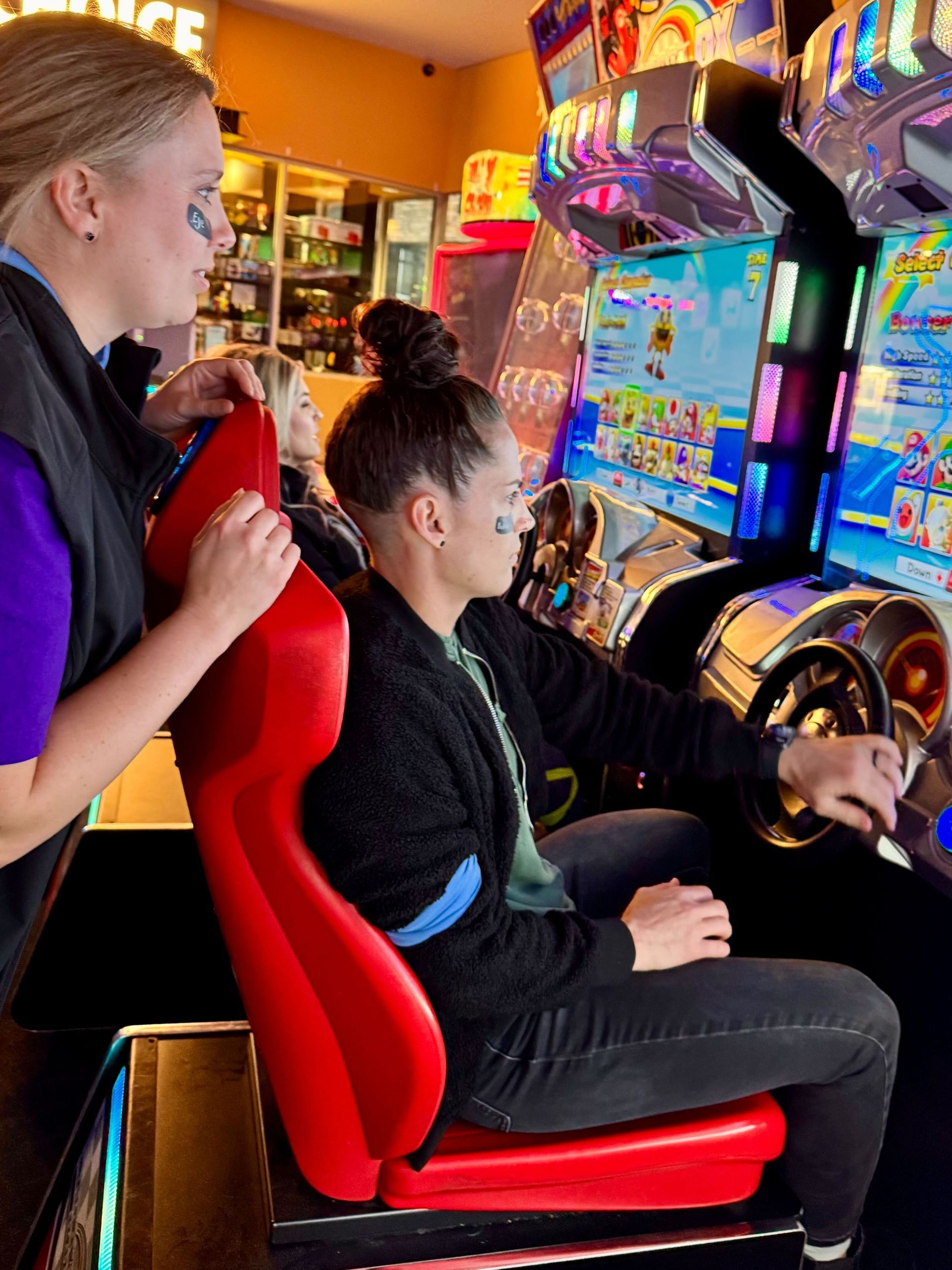 Two women playing a racing arcade game in an arcade. One is steering, the other is observing.