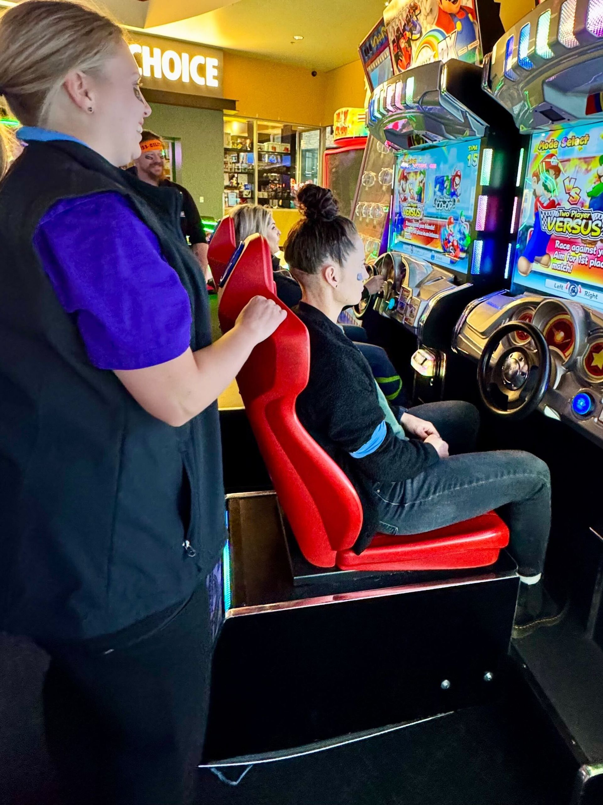 Woman playing a racing arcade game, assisted by an employee in a vest, inside a game arcade.