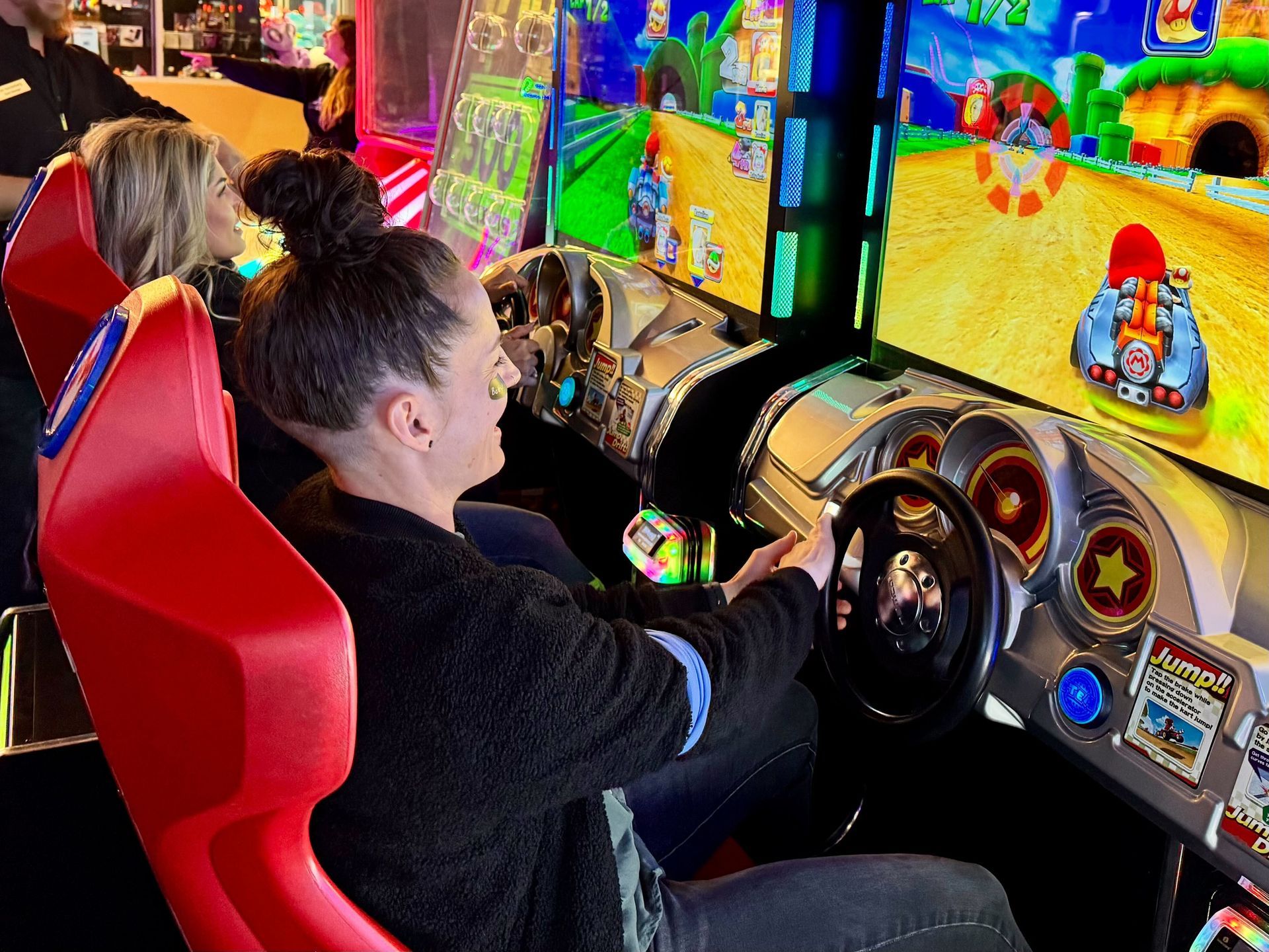 Woman playing racing game in arcade, smiling at the screen, hands on the wheel. Bright colors and arcade lights.
