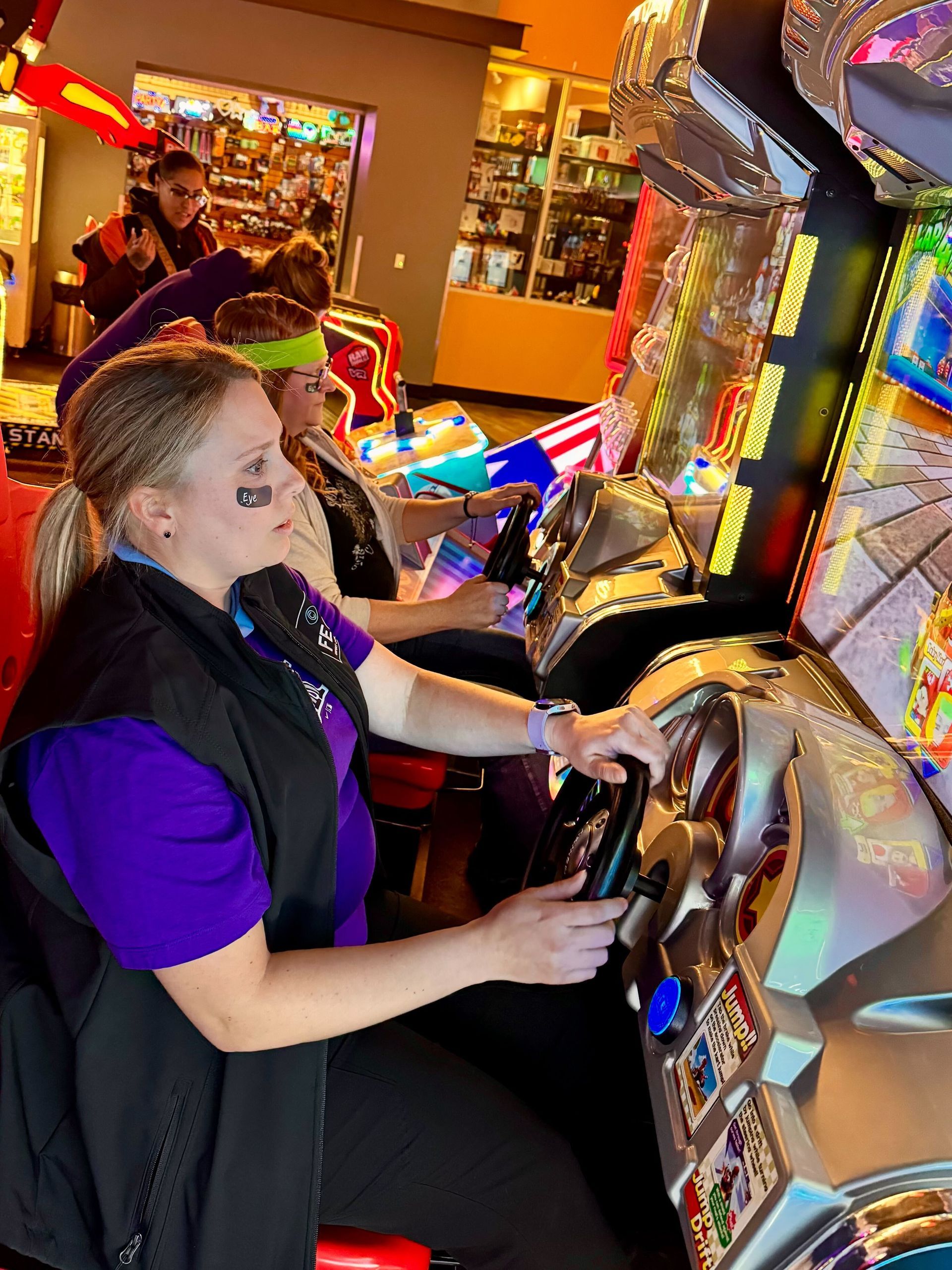 Woman in purple shirt plays arcade game, with others behind her in a brightly lit arcade.