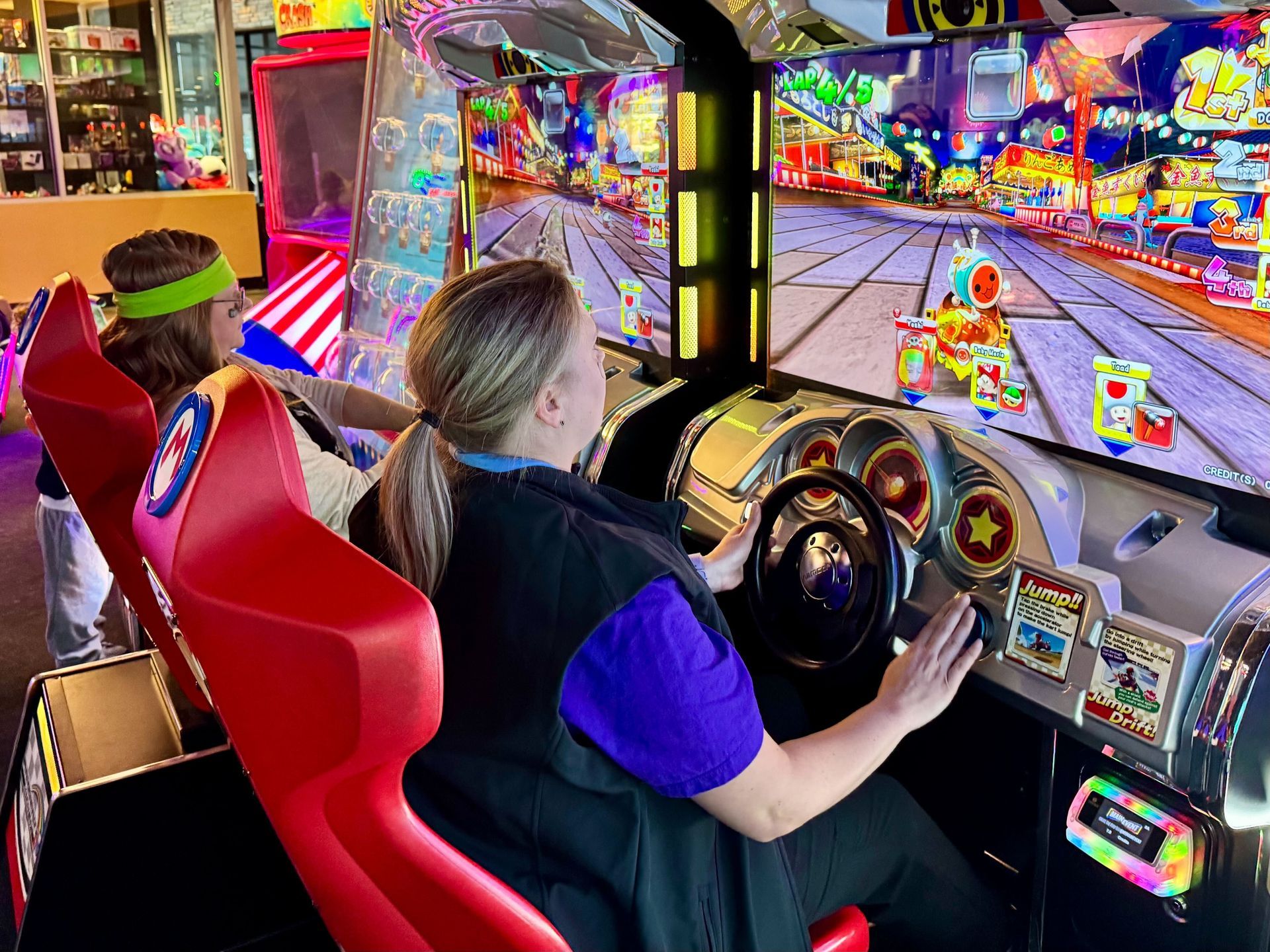Two people playing a colorful racing arcade game. Woman drives, man watches. Bright lights and setting.