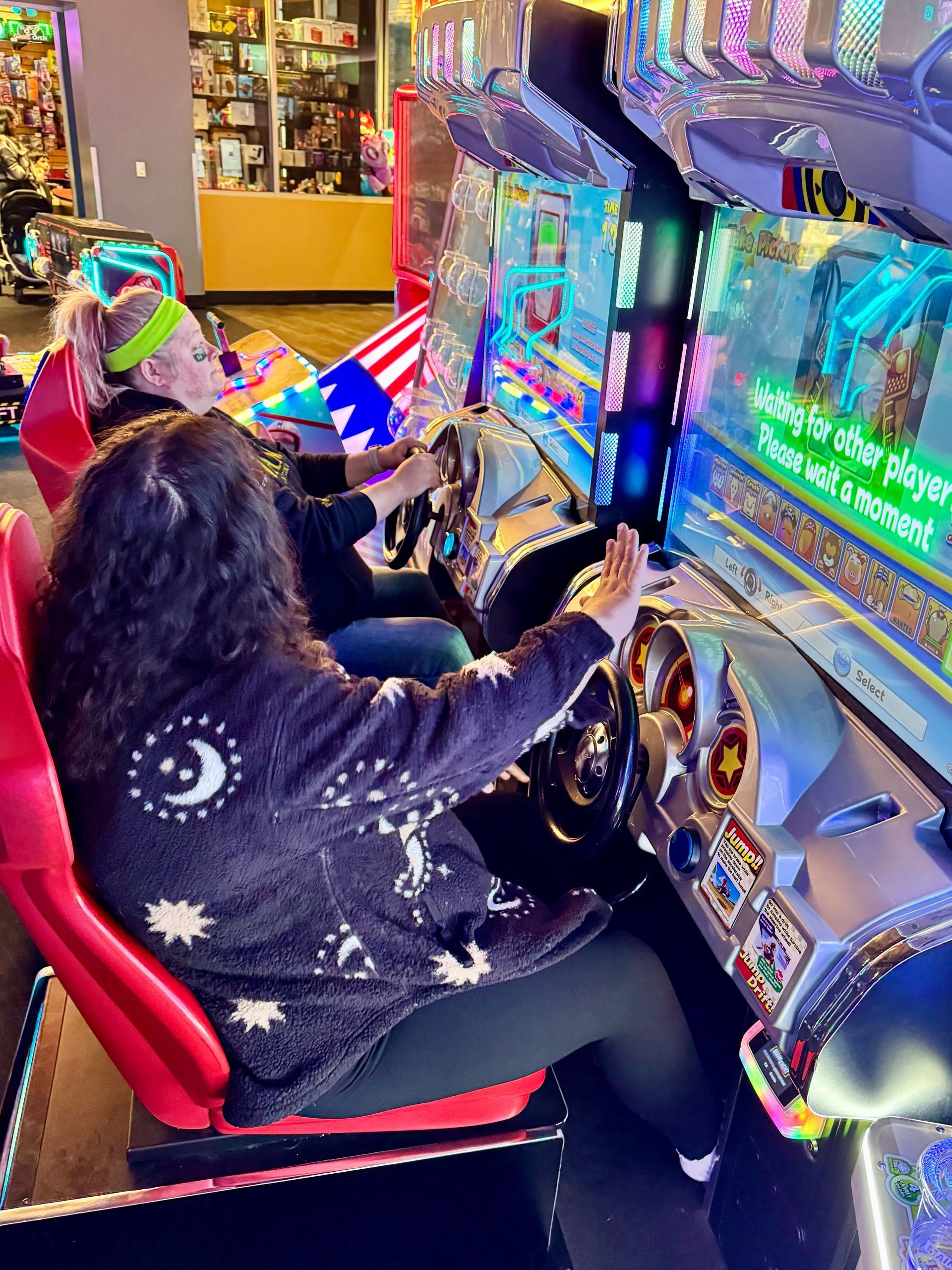 Two people playing a racing arcade game. One wears a black sweater with celestial designs, and the other has a flag headband.