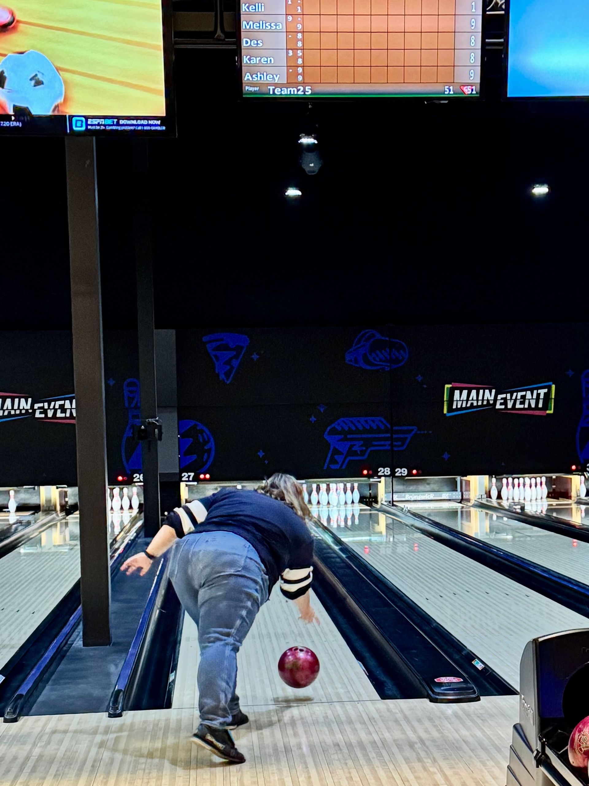 Person bowling at Main Event. They wear a blue shirt, jeans, and are releasing a red bowling ball.