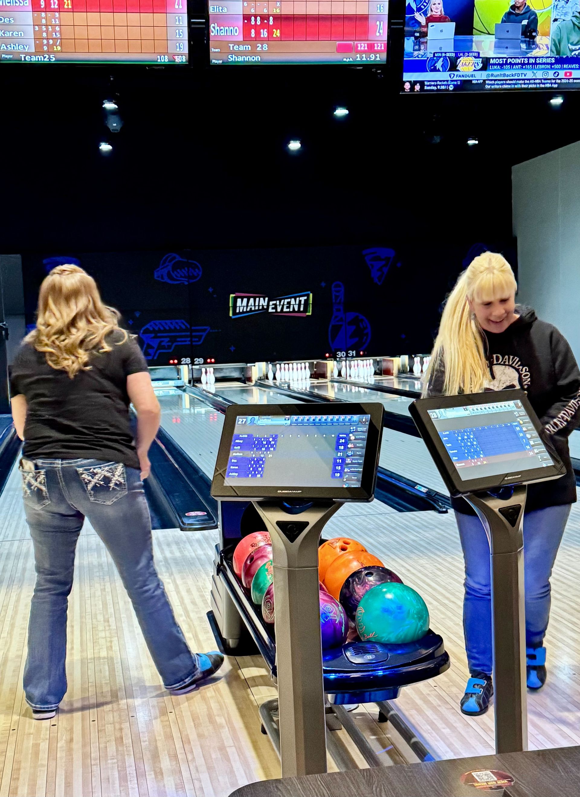 Two women bowling; one in jeans preparing to bowl, the other smiling, next to a bowling ball rack.