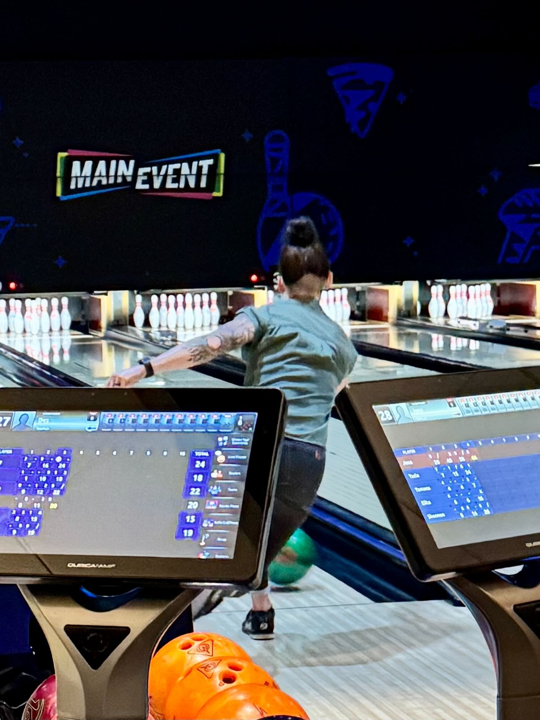 Woman bowling at Main Event; green shirt, arm tattoos, ball rolling down lane.