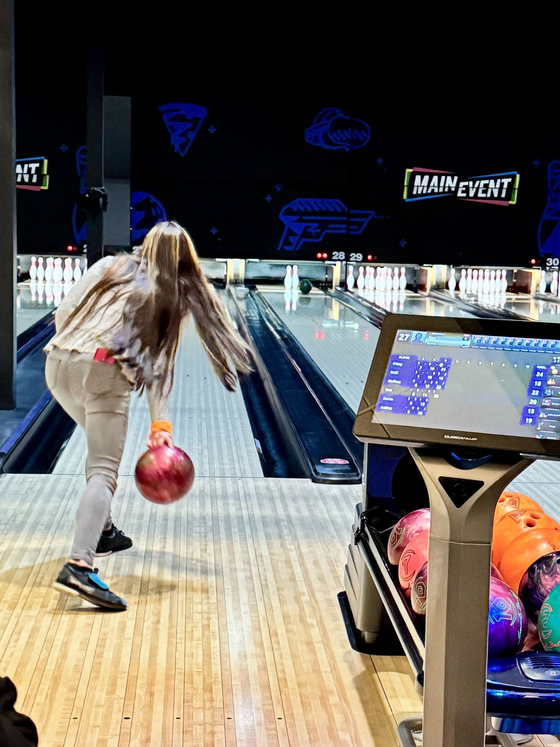 Woman bowling, releasing a red ball. Setting: Main Event bowling alley with digital score display.