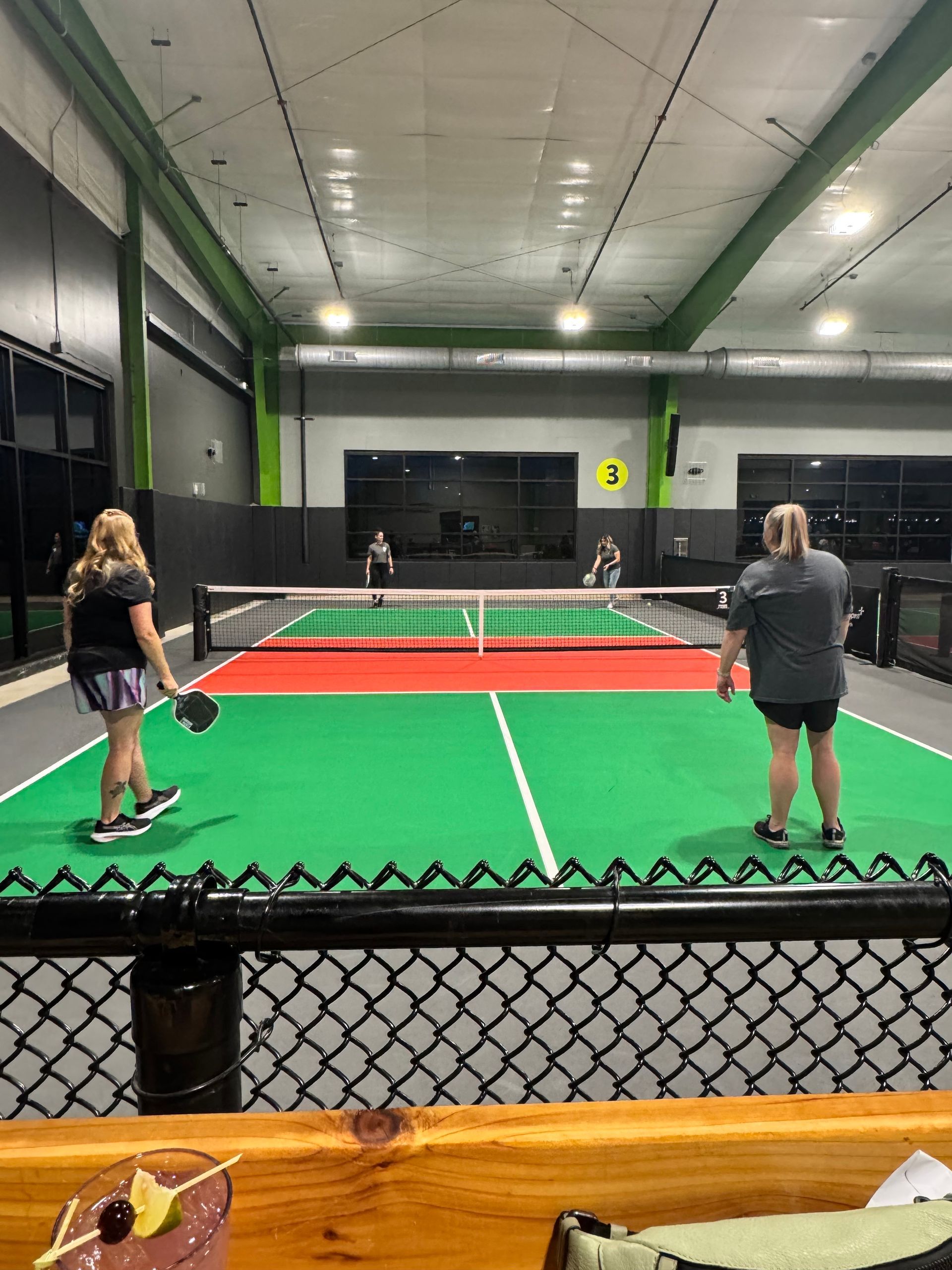 Two women playing pickleball on a green court. One prepares to hit, the other stands ready. Indoor court.