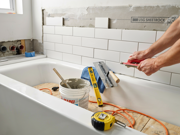 Tiling a bathroom wall above a bathtub, with tools and supplies on the tub edge.