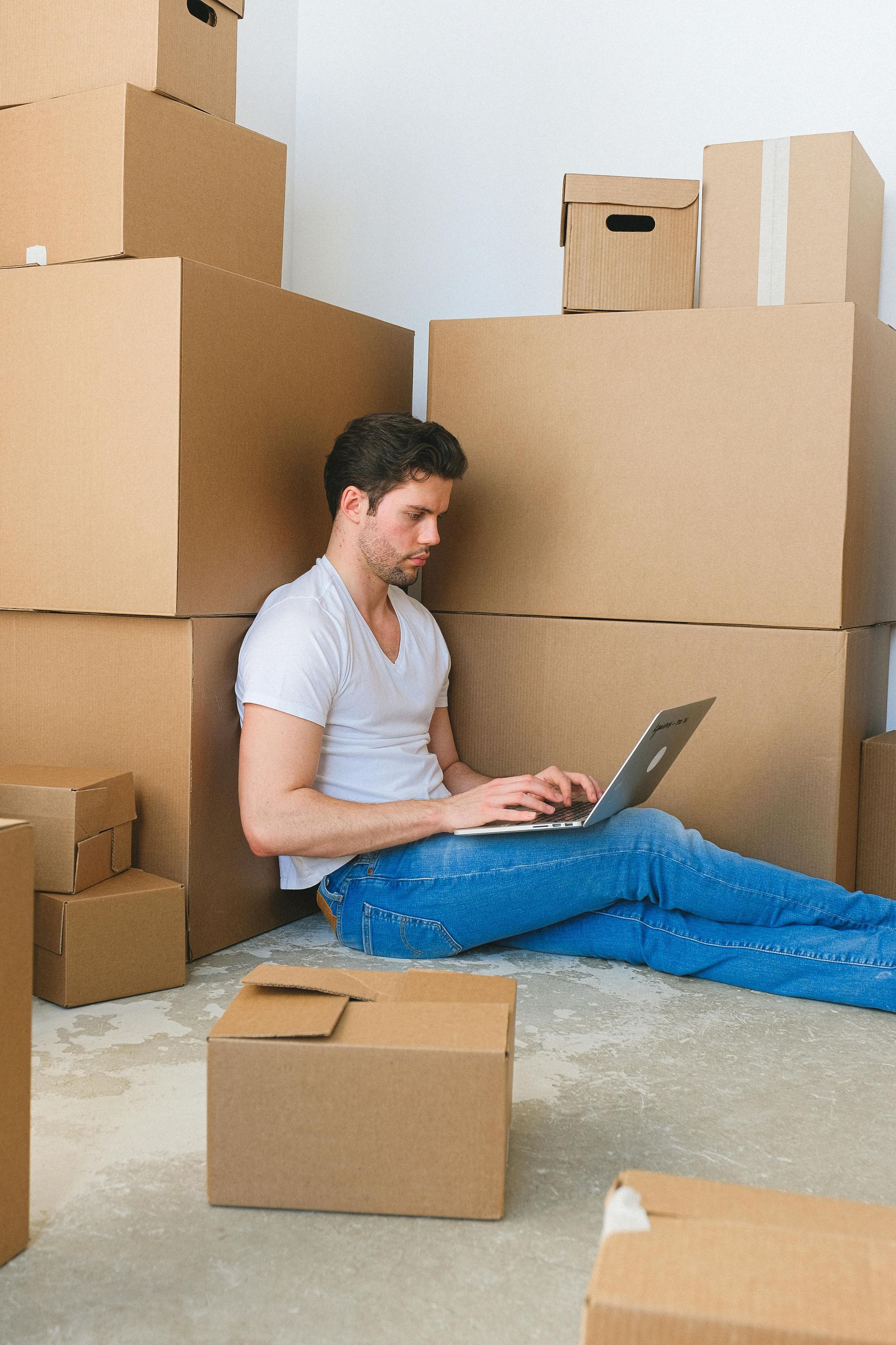 Man sitting on floor, using laptop, surrounded by moving boxes.