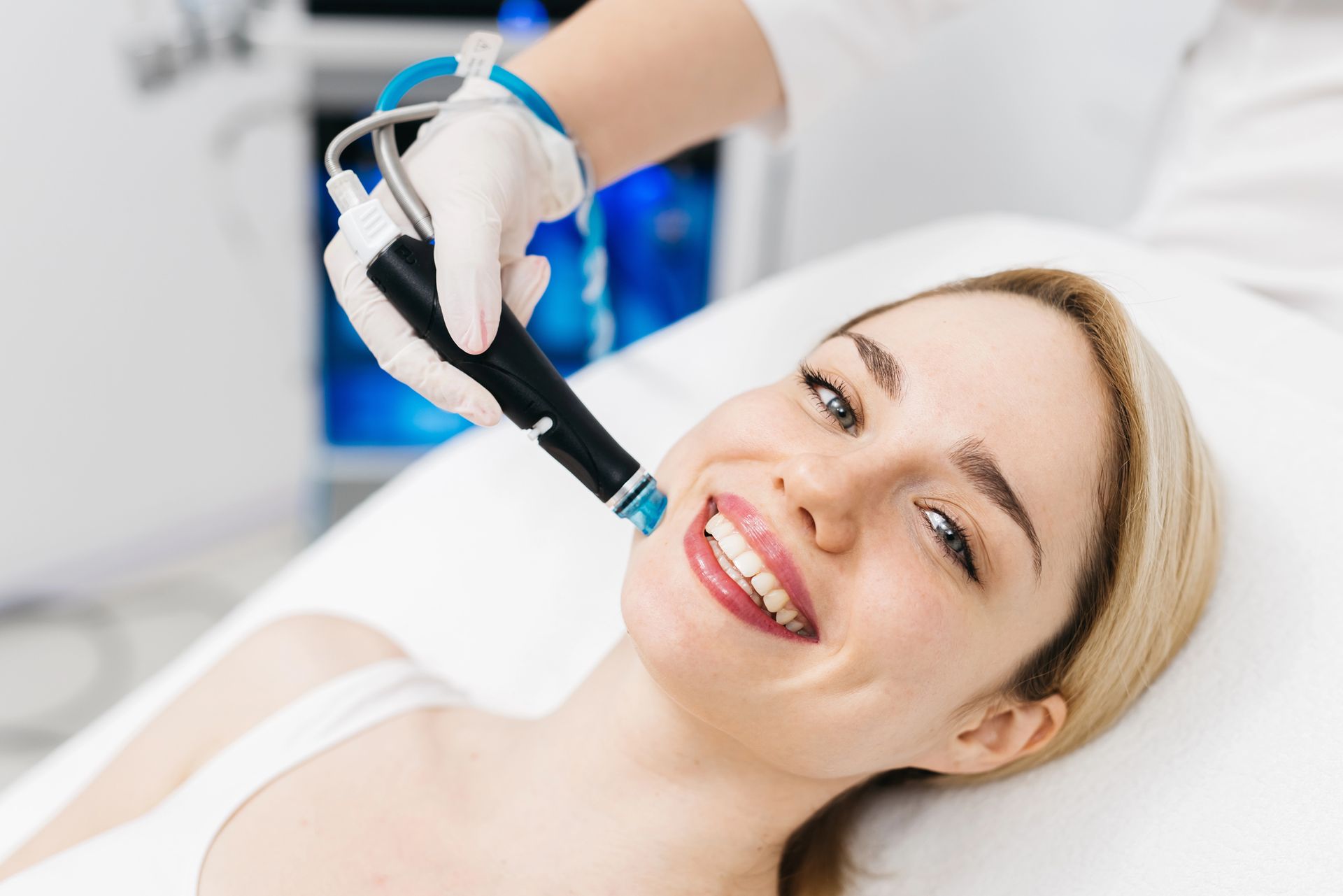 Woman undergoing a facial treatment with a handheld device, smiling on a white surface.