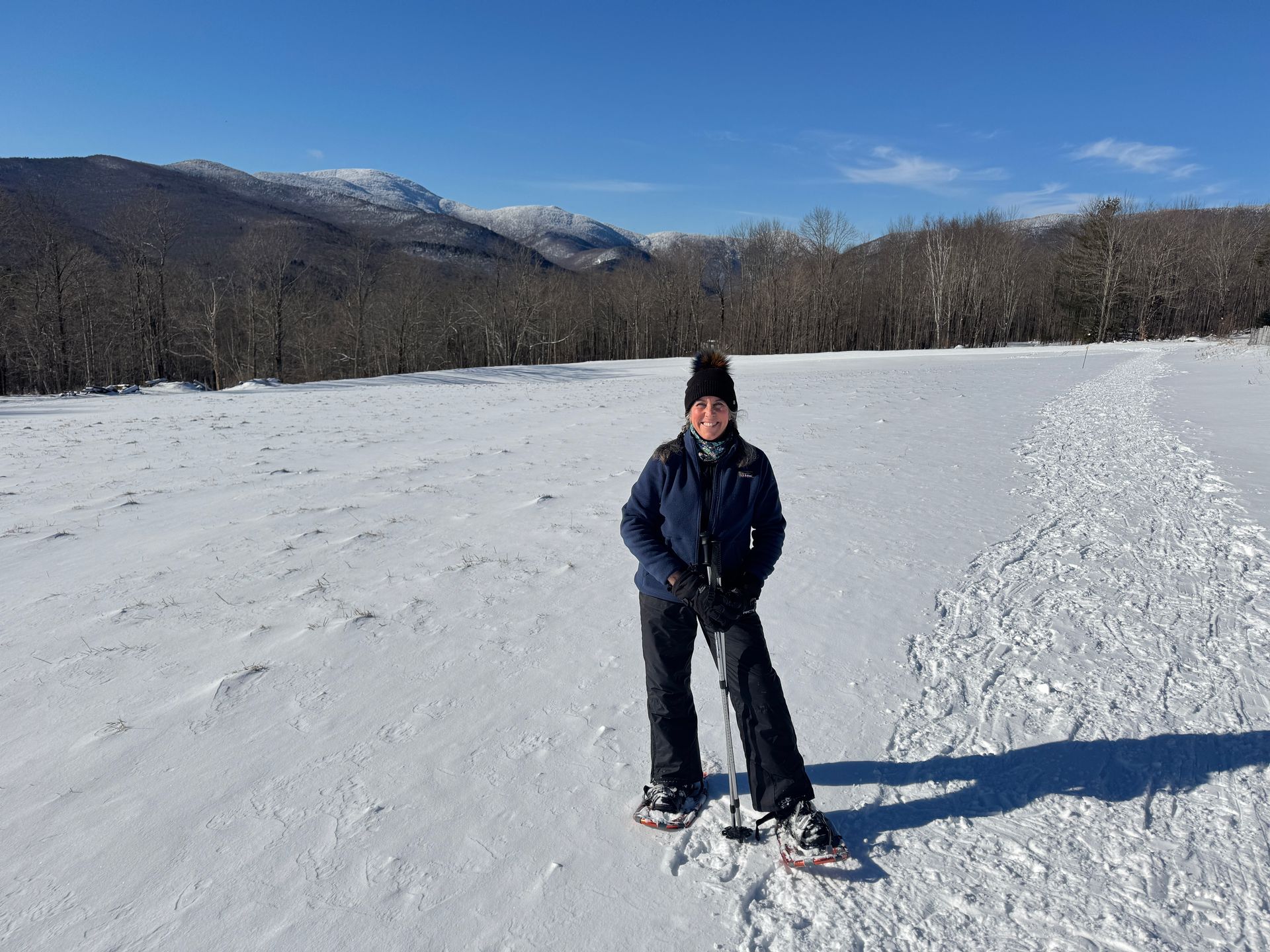 Person snowshoeing in snowy field, mountains in background, blue sky.