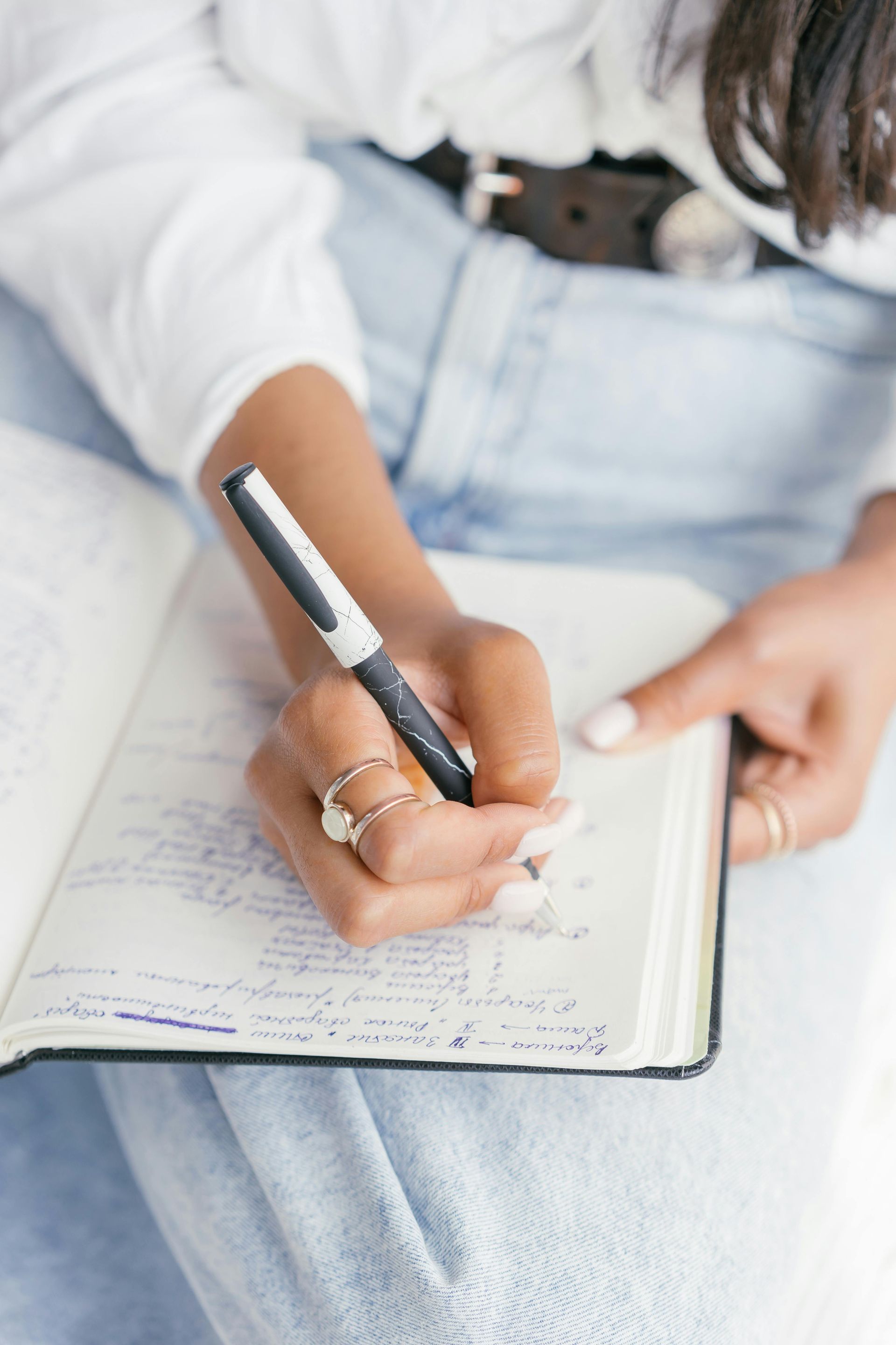 Person writing in a notebook with a black pen. Light blue jeans, white shirt, and rings on fingers.