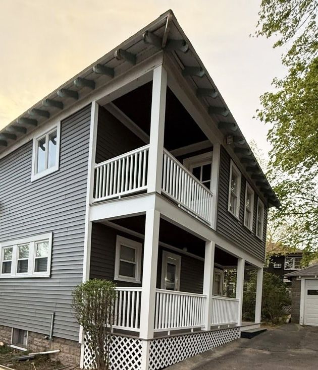 A two-story house exterior with grey siding, white trim, a double-decker front porch with railings, and a lattice base.