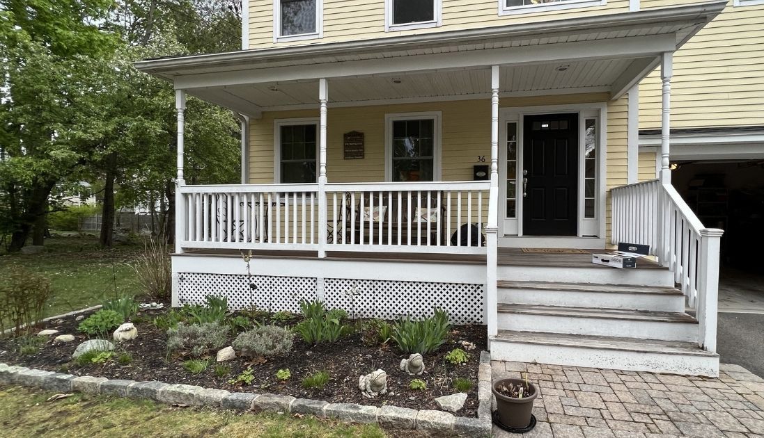 A yellow house with a covered front porch featuring white railings, steps, and a landscaped bed in the front yard.