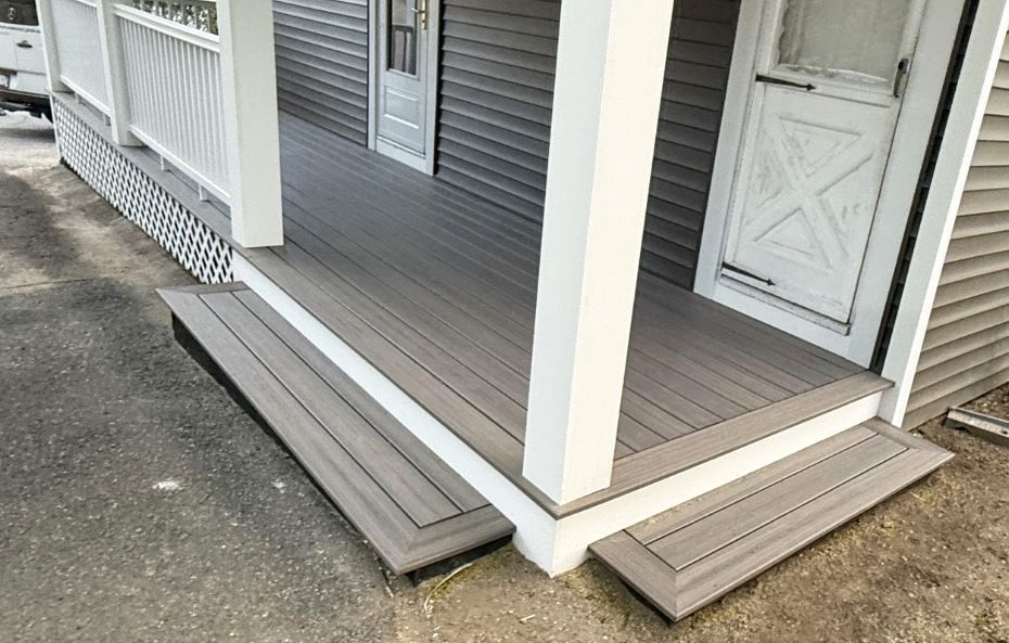 A gray porch with a wooden deck and white trim, featuring two low steps leading up to white doors against gray siding.