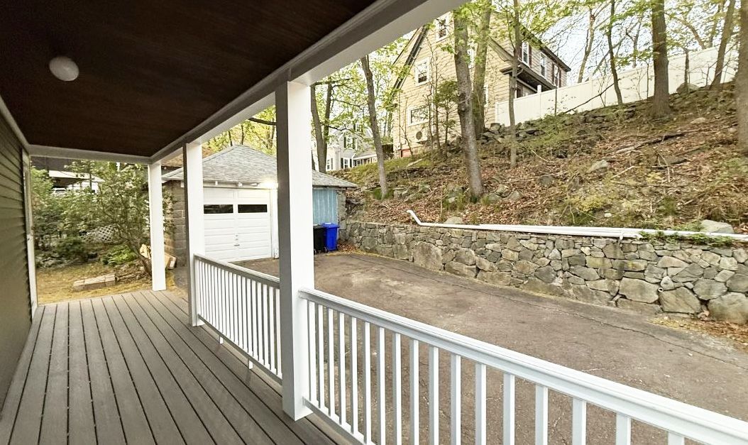 A wooden porch with white railings overlooks a paved driveway and a detached garage next to a stone retaining wall.