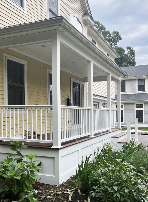 A yellow house with a white front porch, railing, and lattice skirting, surrounded by green plants.