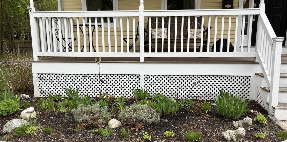 A yellow porch with white railings and lattice skirting, featuring a garden bed with plants and decorative stone figures.