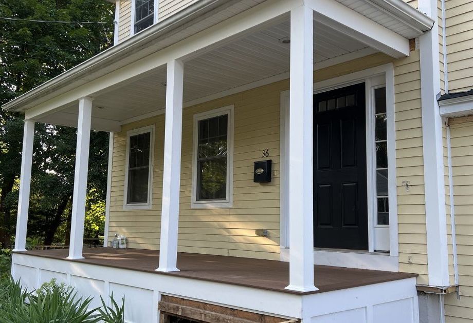 A house with yellow siding and a covered front porch featuring white columns, a dark wooden floor, and a black front door.