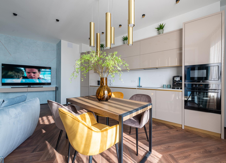 A modern dining and kitchen area with a wooden table, yellow and grey chairs, a centerpiece vase, and sleek cabinetry.