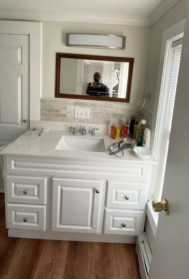 A white bathroom vanity with a marble countertop, sink, mirror, and light fixture in a room with wood-look flooring.