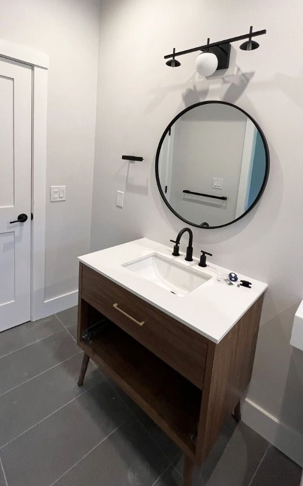 A modern bathroom vanity with a wood-tone base, white countertop, round black mirror, and sleek light fixture.