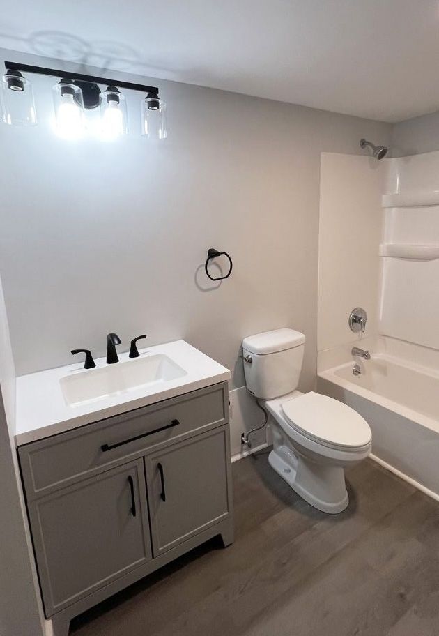 Modern bathroom with a gray vanity, white countertop, toilet, and bathtub with a shower head against light gray walls.