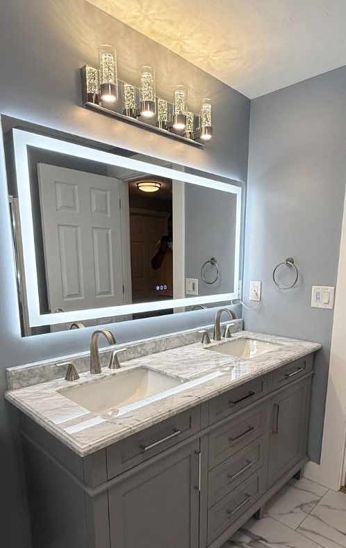 A grey double vanity with a marble countertop, dual sinks, a large rectangular LED mirror, and overhead light fixtures.