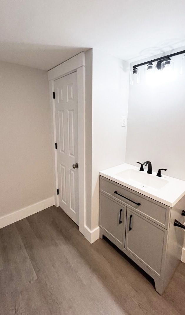 A modern gray vanity with a white countertop and black faucet next to a closed white door in a neutral-toned bathroom.