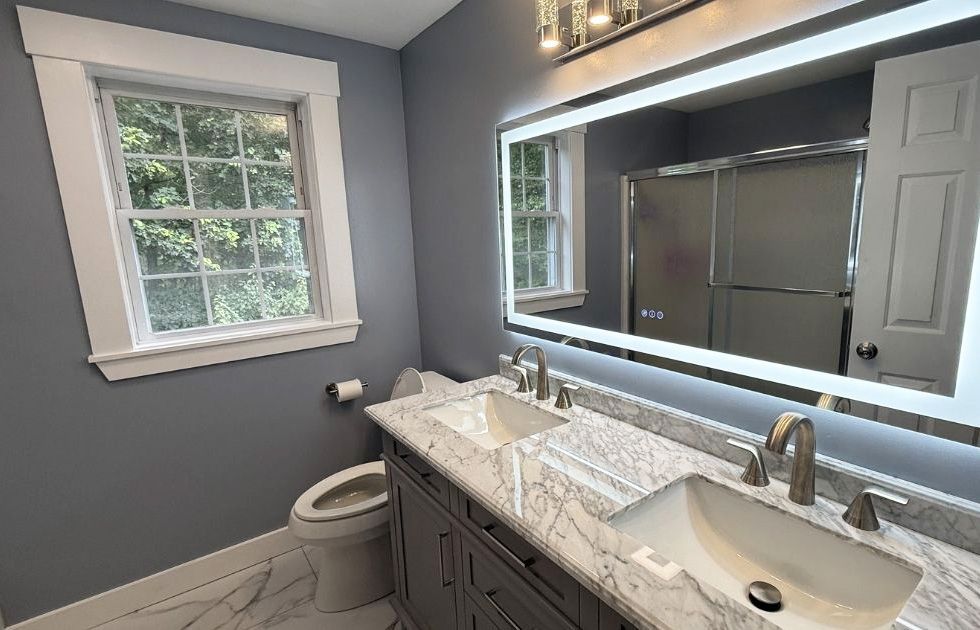 A modern bathroom with blue-grey walls, a double sink marble vanity, and a large, rectangular illuminated LED mirror.