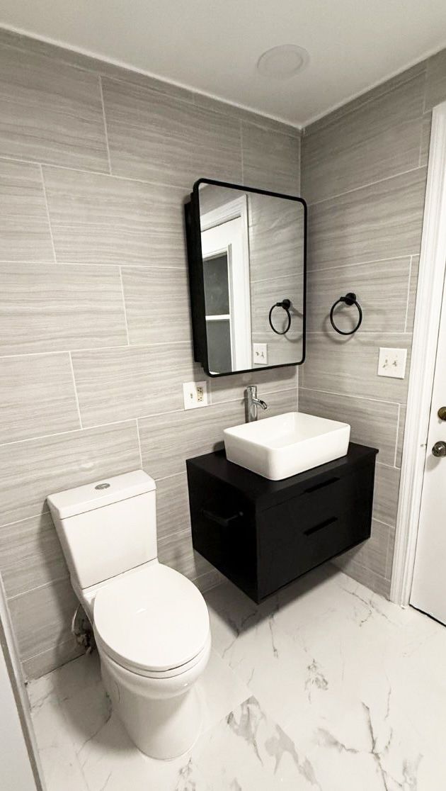 A modern bathroom with a white toilet, black floating vanity with a white vessel sink, and gray horizontal-tiled walls.