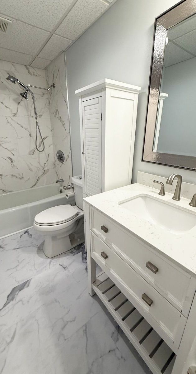 A bathroom with a marble-patterned tub and floor, a white vanity with drawers, a tall storage cabinet, and a wall mirror.