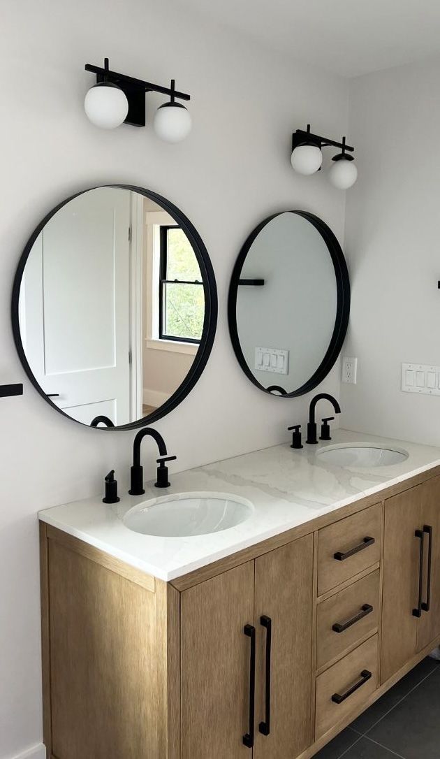 A double bathroom vanity with wood cabinets, white countertops, black faucets, two round mirrors, and black globe lights.