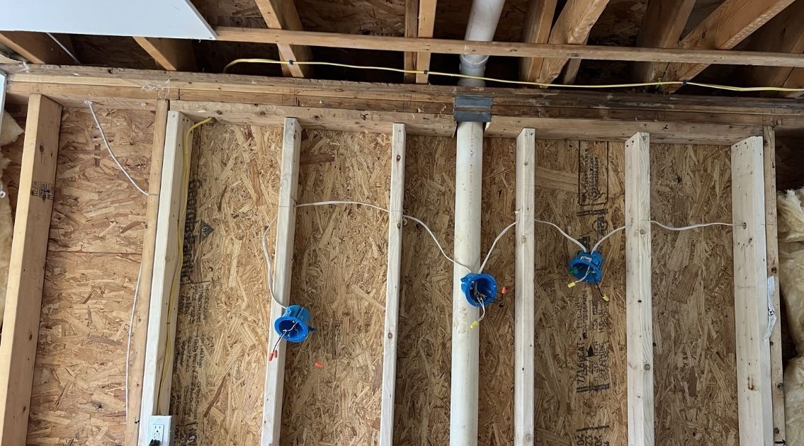 Exposed wall studs with blue electrical boxes, white wiring, and a vertical plumbing pipe in a construction setting.