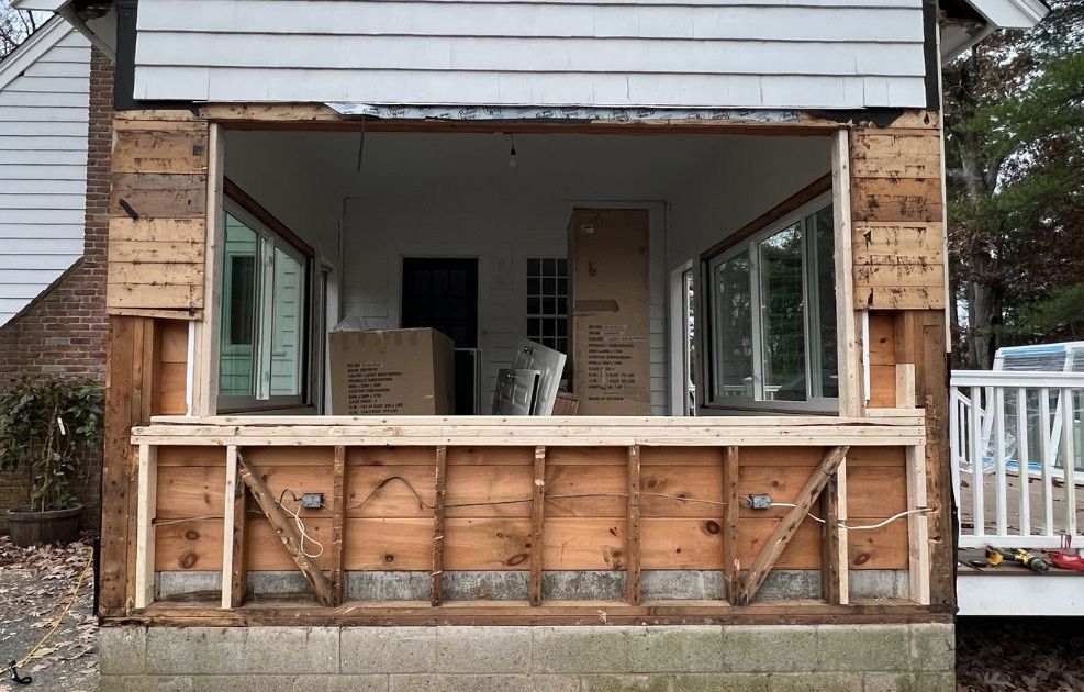 Exterior view of a home renovation featuring an exposed wood-framed wall opening where a large window was removed.