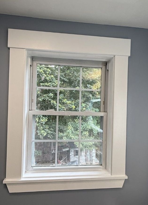A white, wooden window frame with a grid design set against a grey wall with a view of green trees outside.