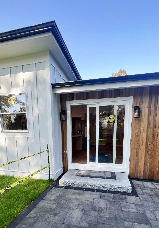 A sliding glass door entrance to a modern home featuring vertical white board-and-batten siding and natural wood paneling.