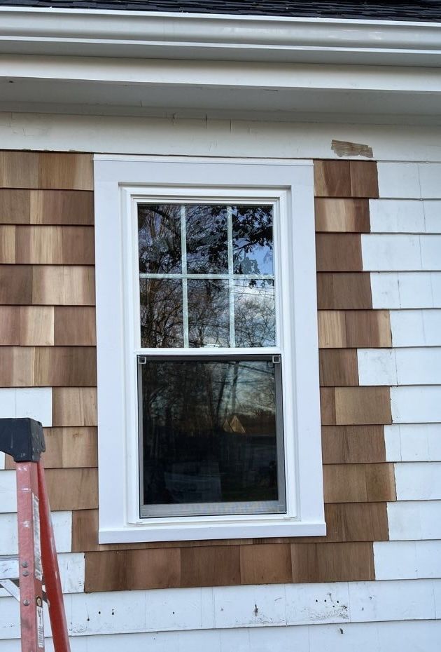 A white-framed window on a house exterior featuring a mix of natural wood and white siding shingles.