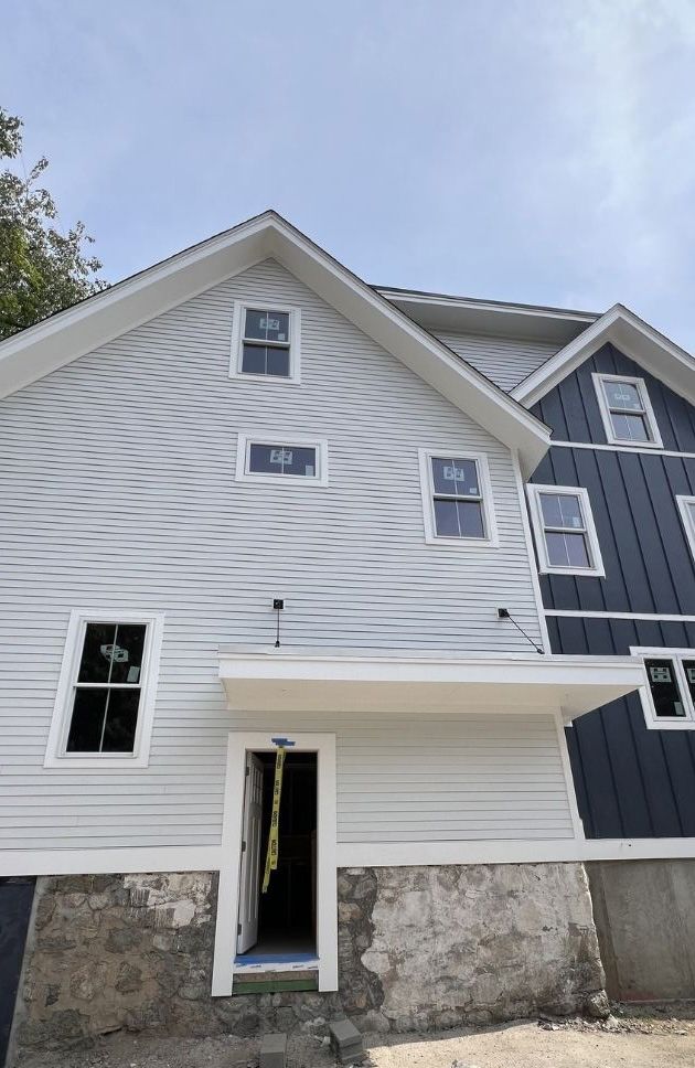 A two-story house under construction with grey horizontal siding, a dark blue vertical-siding section, and stone foundation.