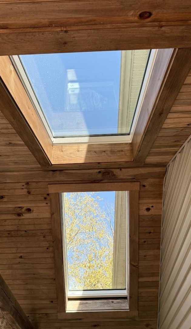 Two skylights set into a wooden ceiling, revealing blue sky and autumn tree branches through the glass.