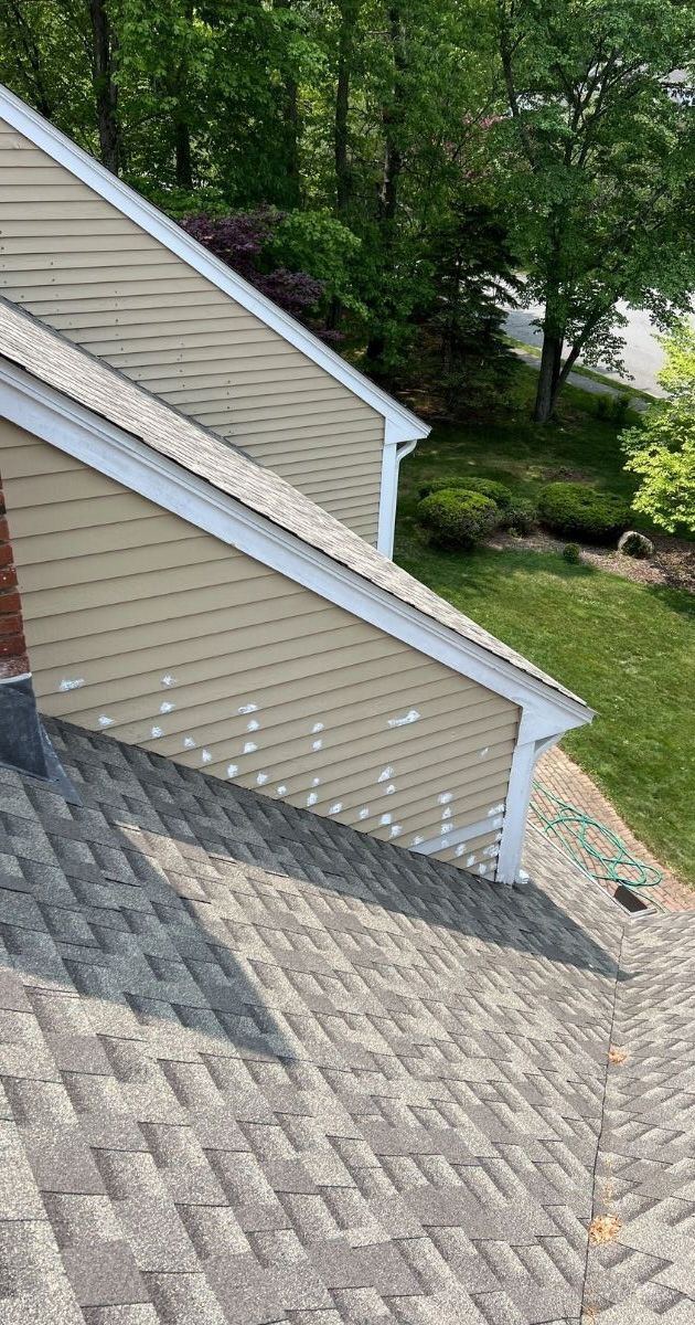 A high-angle view of a house roof with light and dark gray shingles, meeting a tan-sided wall with white trim.